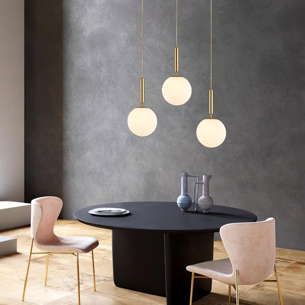 Three brass-and-white Clérachic sphere pendants hang low above a black oval dining table with pink chairs.