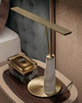 The Clérachic table lamp's white marble base and bronze bar sit on a wooden nightstand beside a tufted grey-green headboard, unlit.