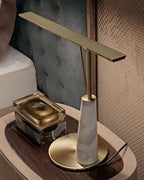 The Clérachic table lamp's white marble base and bronze bar sit on a wooden nightstand beside a tufted grey-green headboard, unlit.