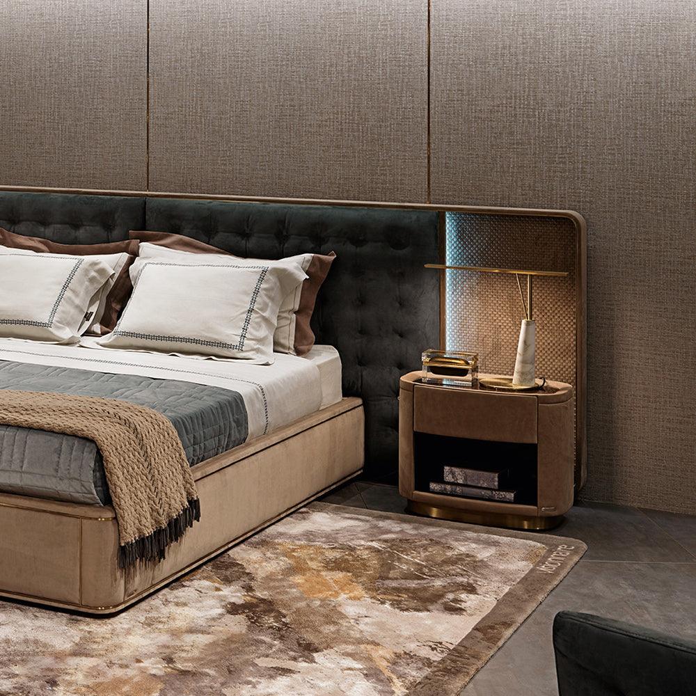 Lit on a bedroom nightstand, the Clérachic gold table lamp with its marble cone base sits beside a bed dressed in taupe and grey linens.