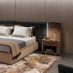 Lit on a bedroom nightstand, the Clérachic gold table lamp with its marble cone base sits beside a bed dressed in taupe and grey linens.