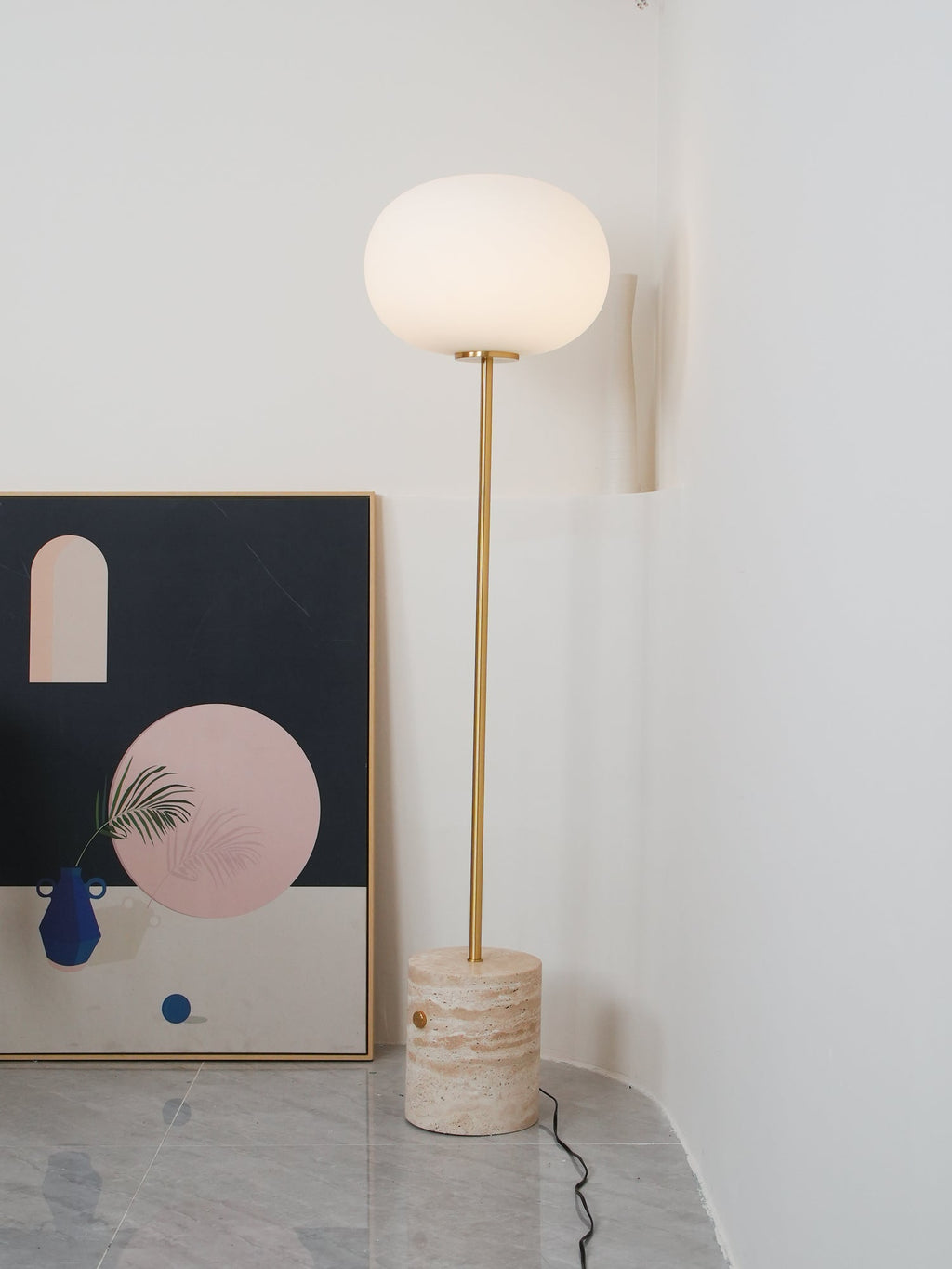 The Clérachic floor lamp lit in a plain white corner, with an abstract framed print leaning against the wall on tiled floor.