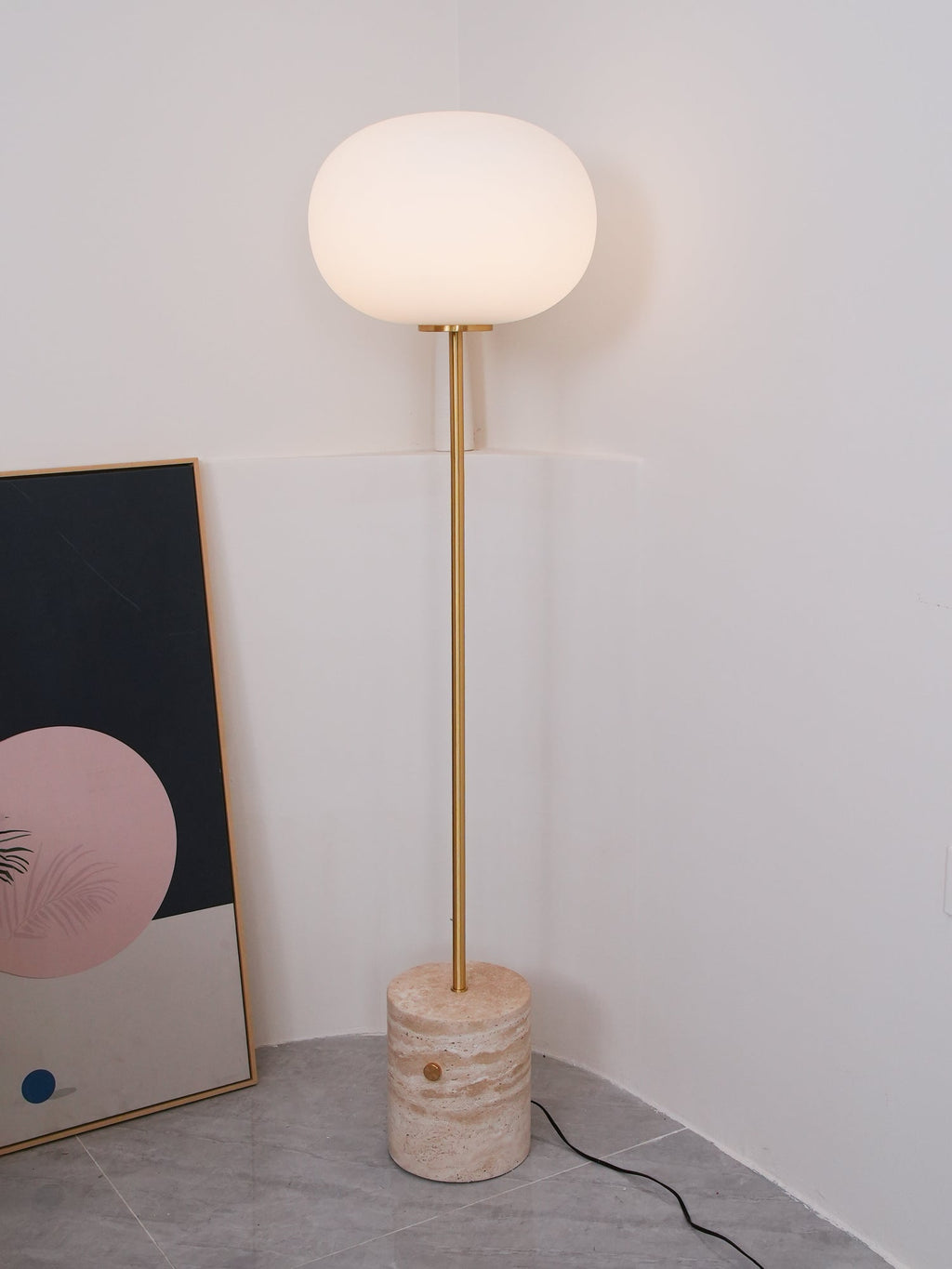 The Clérachic floor lamp's frosted globe glows warmly in a plain white corner beside a pink and navy abstract print.