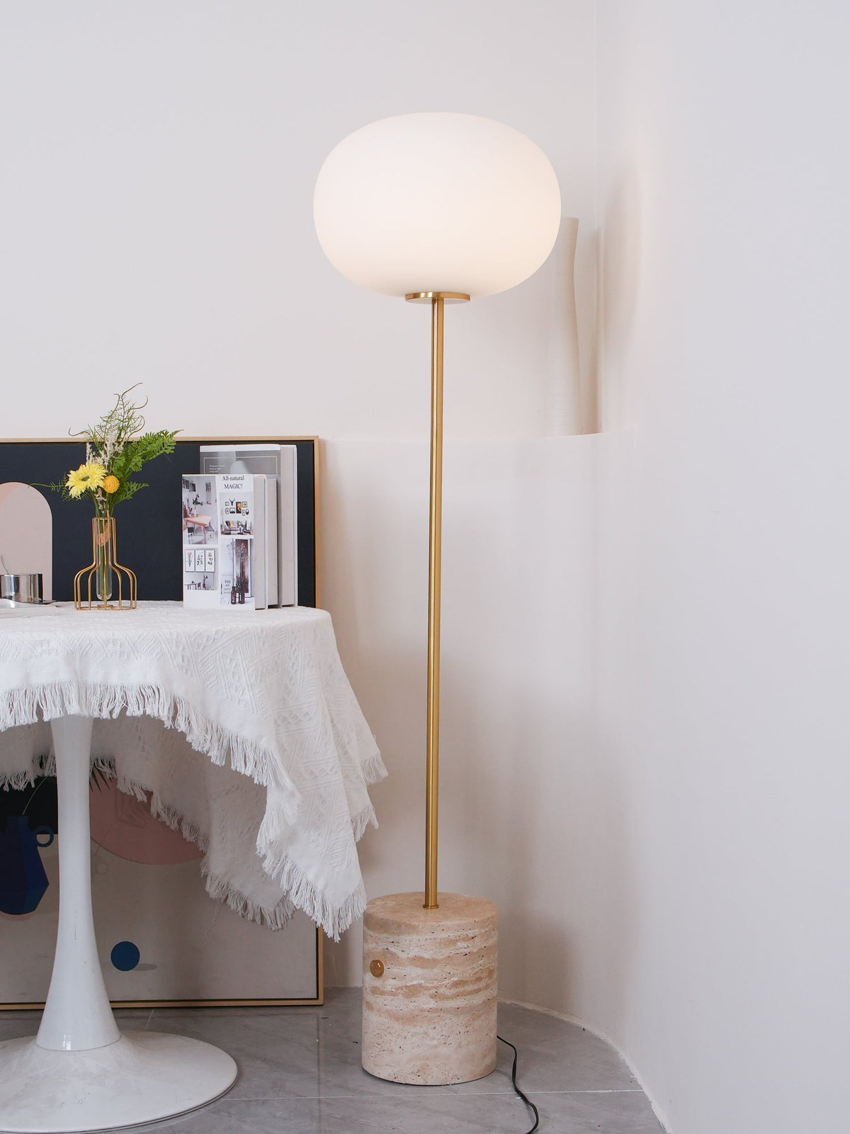 The Clérachic floor lamp stands near a fringed white tablecloth set with fresh flowers and magazines, on a tiled floor.