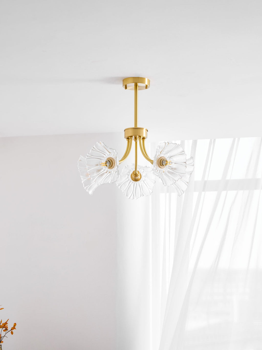 A small dried flower sprig sits below this Clérachic chandelier, hung near a sheer curtain in a white room.