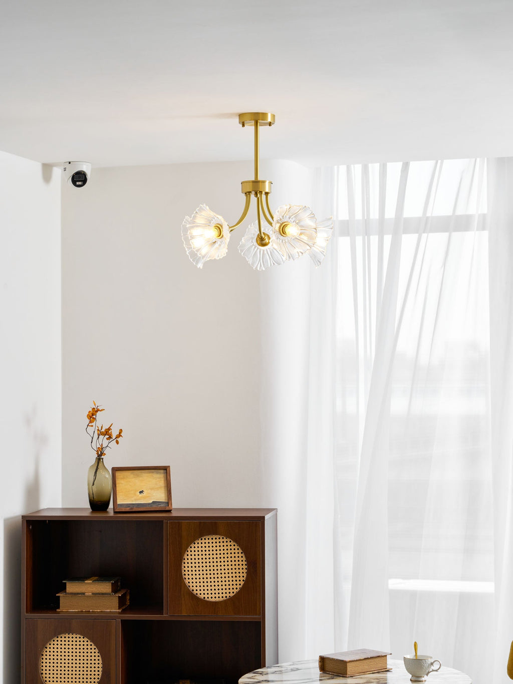 A rattan-fronted cabinet and a vase of dried flowers sit beneath this Clérachic chandelier, hung beside a sheer curtain.
