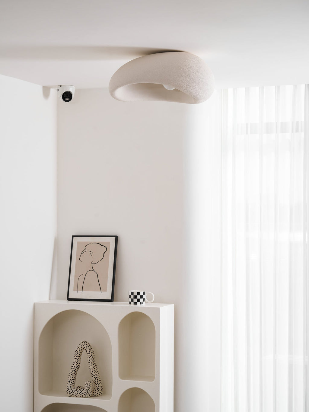 In its white texture finish, this Clérachic ceiling light sits lit near a sheer curtain above a framed sketch and a mug.