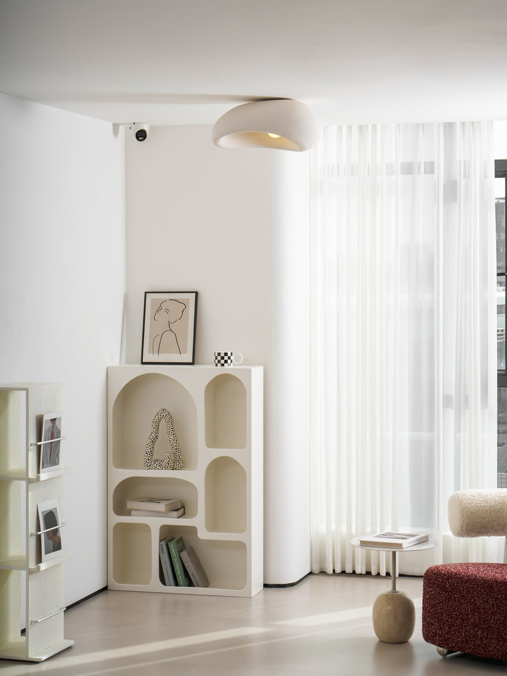 This Clérachic ceiling light lights a corner with a curved bookshelf, sheer curtains and a red ottoman.