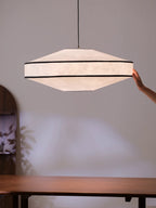 A hand reaches to adjust this Clérachic pendant's cord, its faceted paper shade glowing above a wood table.