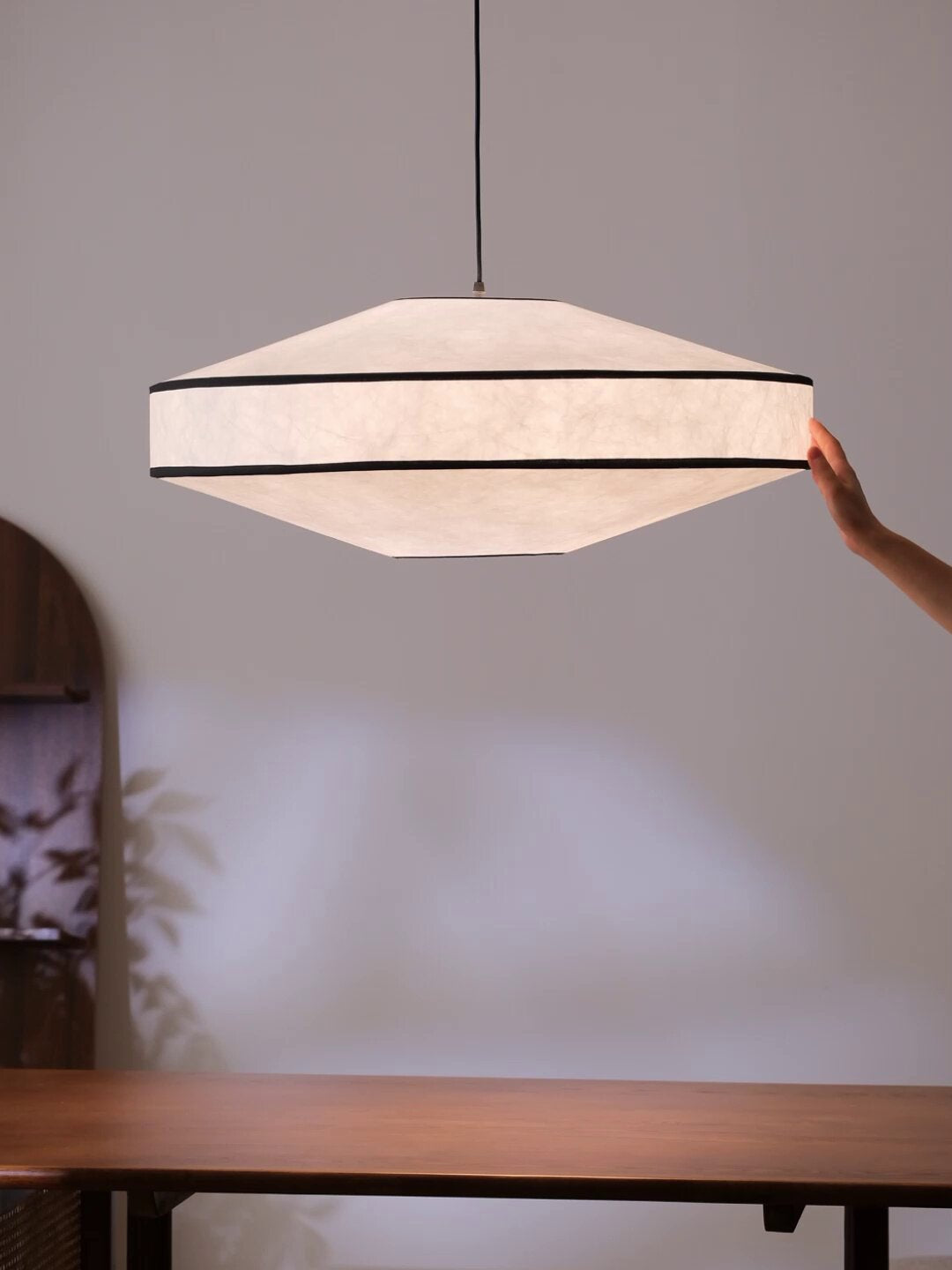 A hand reaches to adjust this Clérachic pendant's cord, its faceted paper shade glowing above a wood table.