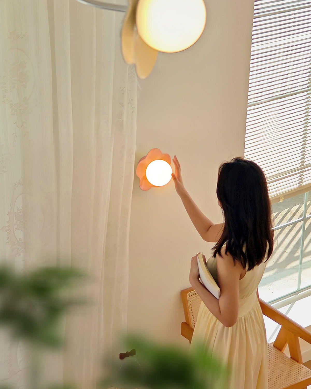 A woman in a yellow dress reaches up to switch on this Clérachic wall light in pink, beside a sheer curtain.