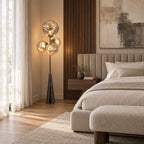 Clérachic floor lamp lit beside a channel-tufted headboard, its smoke-glass globes glowing warmly against sheer bedroom curtains.