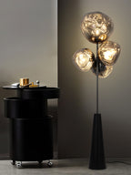 Clérachic floor lamp lit beside a black drinks trolley in a dark, moody room, its smoke-glass globes glowing against a charcoal wall.