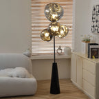 Striped window-blind light falls across a Clérachic floor lamp, its smoke-glass globes glowing beside a cream sideboard on a herringbone floor.