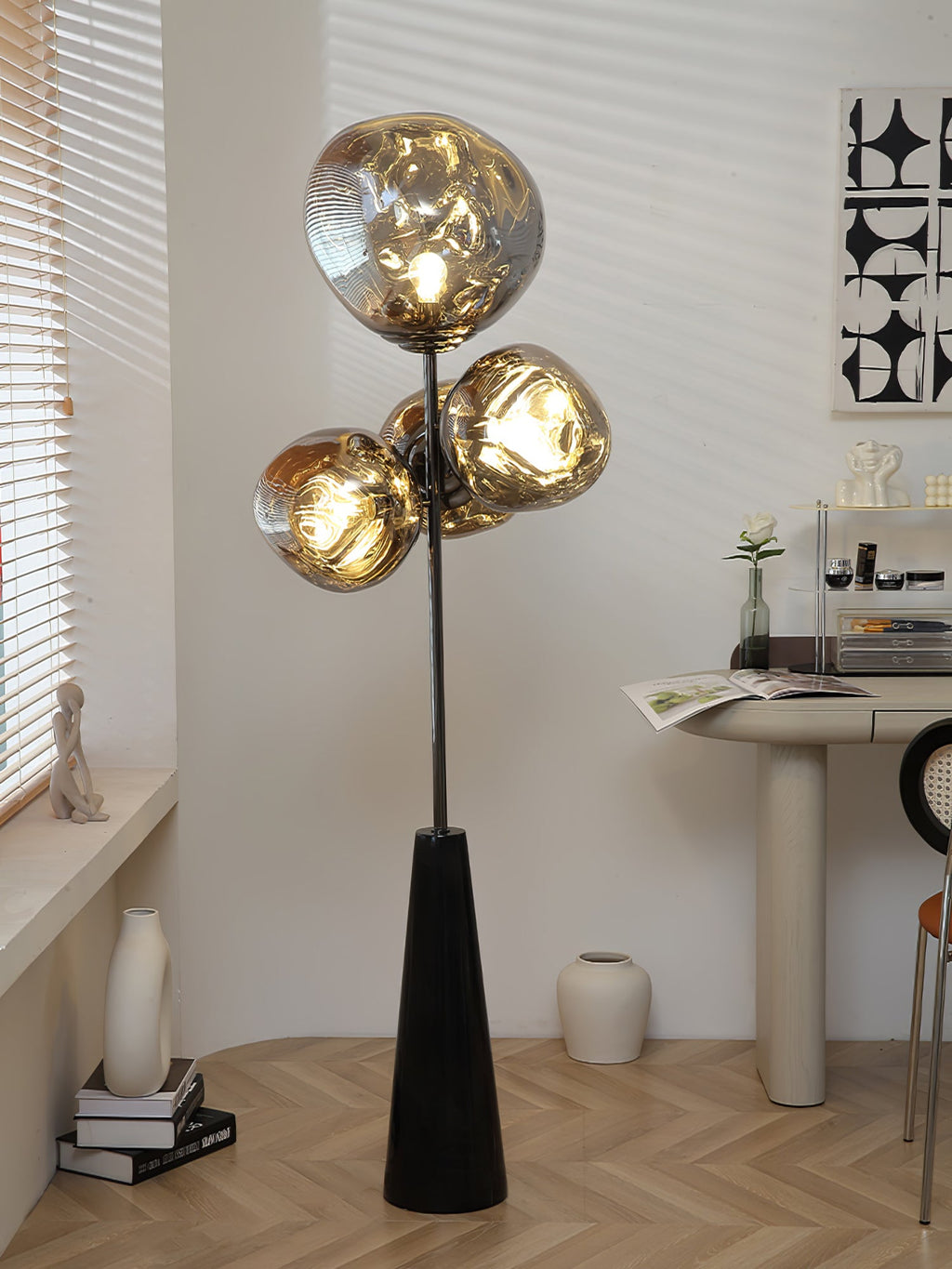 Clérachic floor lamp lit beside a pedestal table, small vase and figurine, blind-slat shadows crossing the herringbone floor behind it.