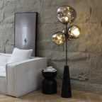Clérachic floor lamp lit against a rough stone accent wall, its marbled glass globes glowing beside a white sofa and round black side table.