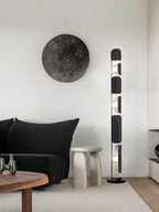 In a black-toned living room, the Clérachic floor lamp glows near a round stone wall piece, a black sofa and a pale stone stool.