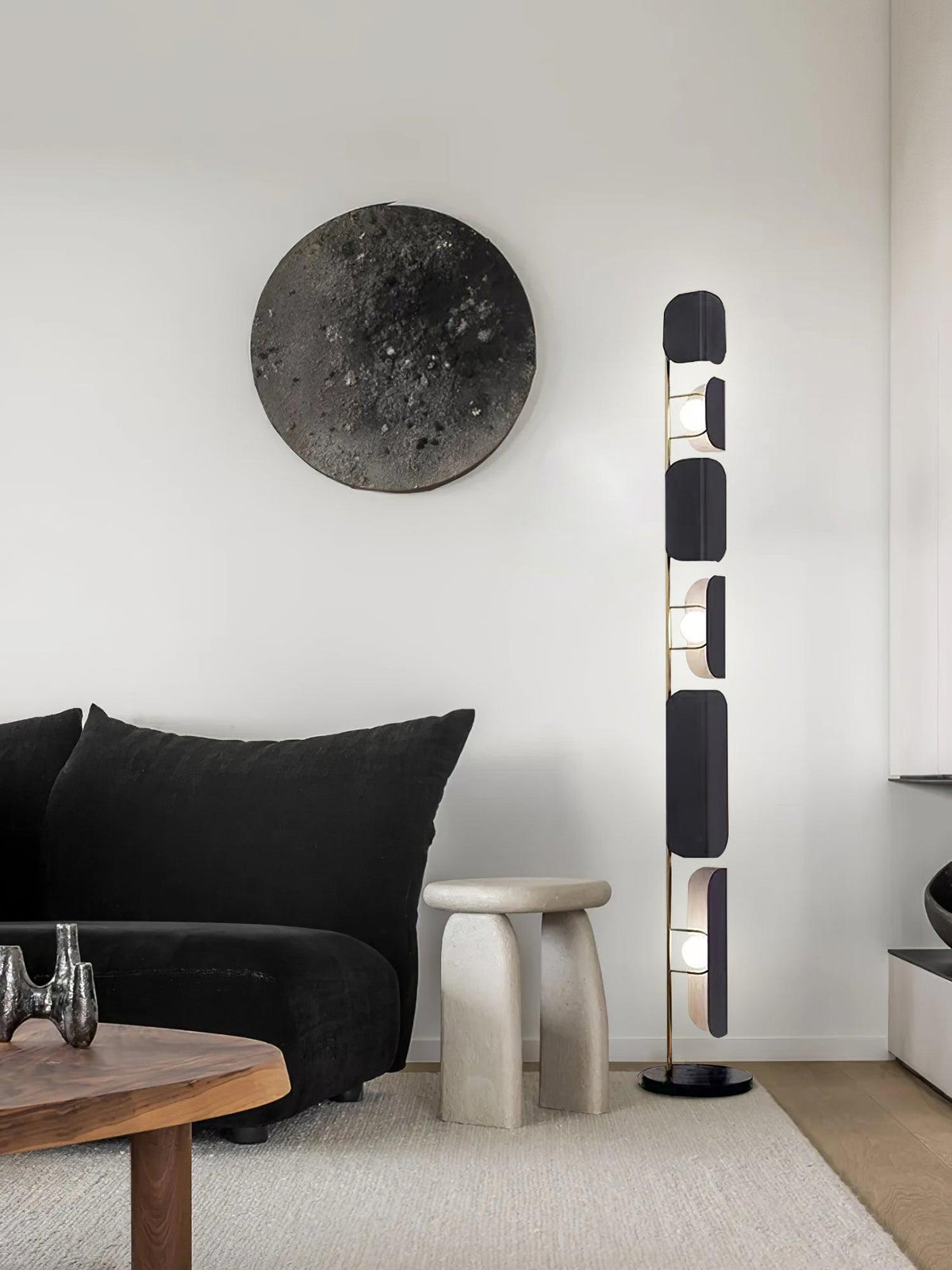 In a black-toned living room, the Clérachic floor lamp glows near a round stone wall piece, a black sofa and a pale stone stool.