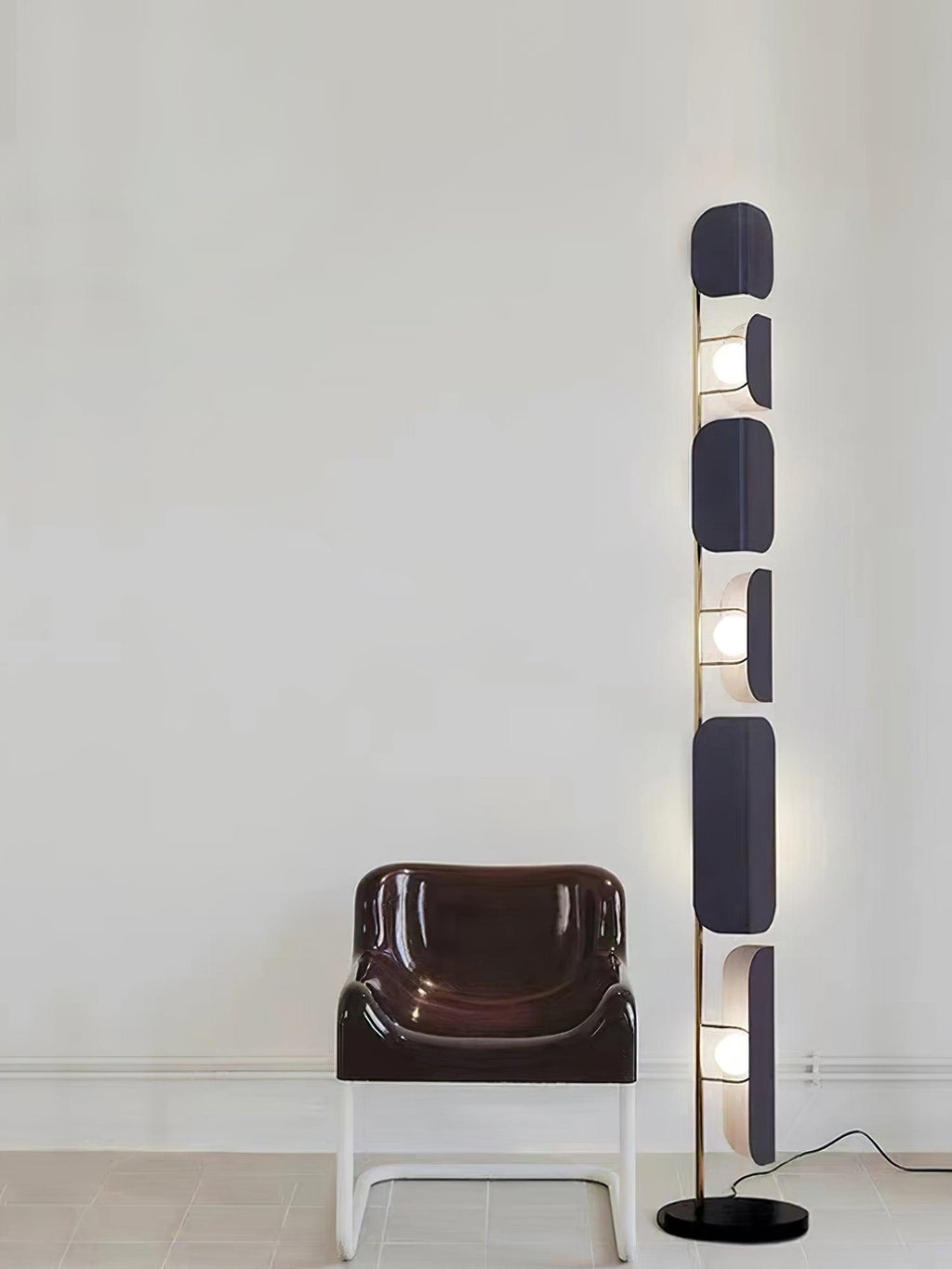 The Clérachic floor lamp lit beside a curved brown leather chair on a pale tiled floor and plain white wall.