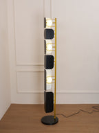 Closer view of the Clérachic floor lamp's black glass panels and gold frame against a plain wall, its cord trailing across the wood floor.