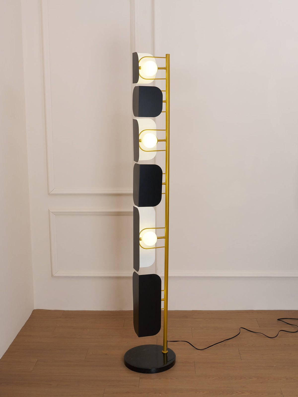 Closer view of the Clérachic floor lamp's black glass panels and gold frame against a plain wall, its cord trailing across the wood floor.