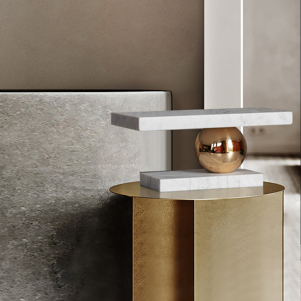 Clérachic white marble table lamp perched on a brushed-gold cylindrical nightstand beside a bed.