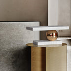 Clérachic white marble table lamp perched on a brushed-gold cylindrical nightstand beside a bed.