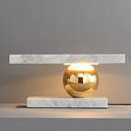 Clérachic white marble table lamp glowing on a grey tabletop, shot close to show the brass sphere and cord.