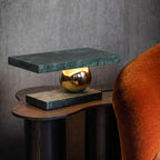 Clérachic green marble table lamp glowing on a wooden side table beside a burnt-orange velvet armchair, seen from a lower angle.