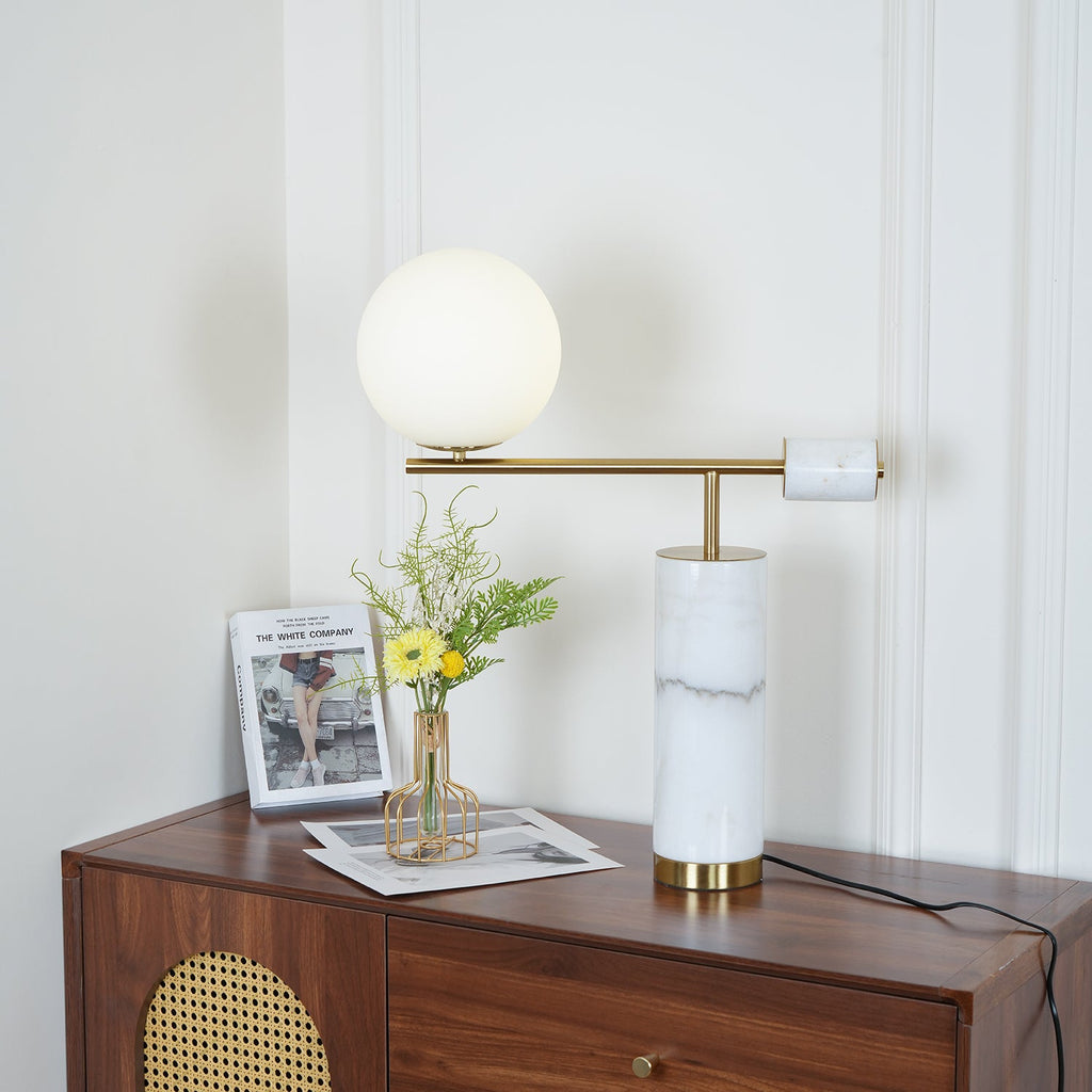 The white marble Clérachic lamp sits lit on a cane-panelled dresser beside a magazine and a small vase of yellow flowers.