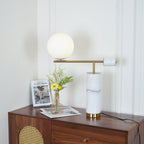 The white marble Clérachic lamp sits lit on a cane-panelled dresser beside a magazine and a small vase of yellow flowers.
