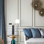 In a green-panelled living room, the lit Clérachic lamp stands on a teal-based side table beside a patterned-cushion sofa.