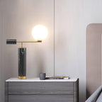 The lit Clérachic lamp sits on a grey wood dresser with a small mug and book, a blush pink headboard behind it.