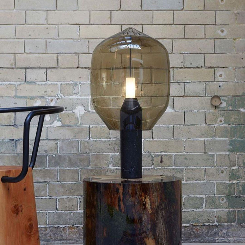 A smoked-glass Clérachic lamp on its black base, standing outdoors on a wood log slab against an old brick wall.