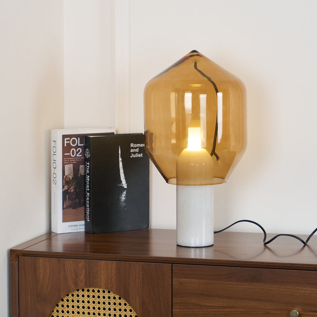 Clérachic's amber-glass lamp on a white marble base, lit beside two stacked books on a walnut credenza, cord trailing to the plug.