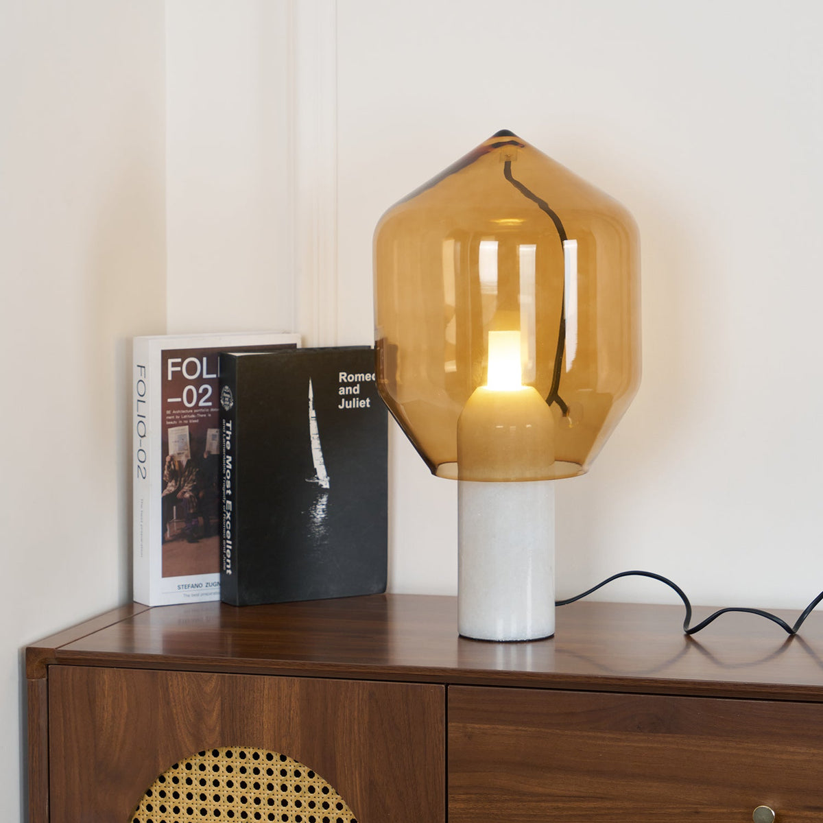 Clérachic's amber-glass lamp on a white marble base, lit beside two stacked books on a walnut credenza, cord trailing to the plug.