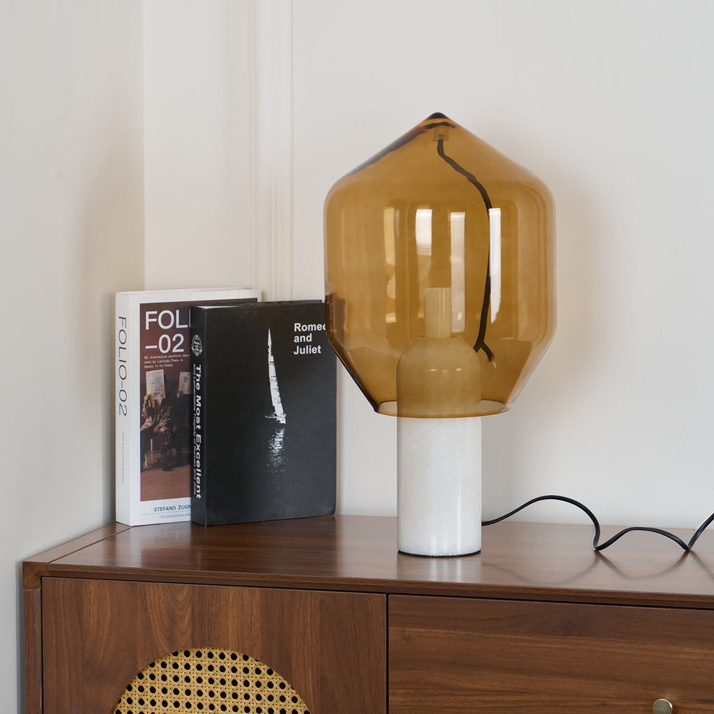 Clérachic's amber-glass lamp on white marble, lit on a walnut credenza beside books and a cane-fronted cabinet.