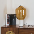 Clérachic's amber-glass lamp on white marble, lit on a walnut credenza beside books and a cane-fronted cabinet.