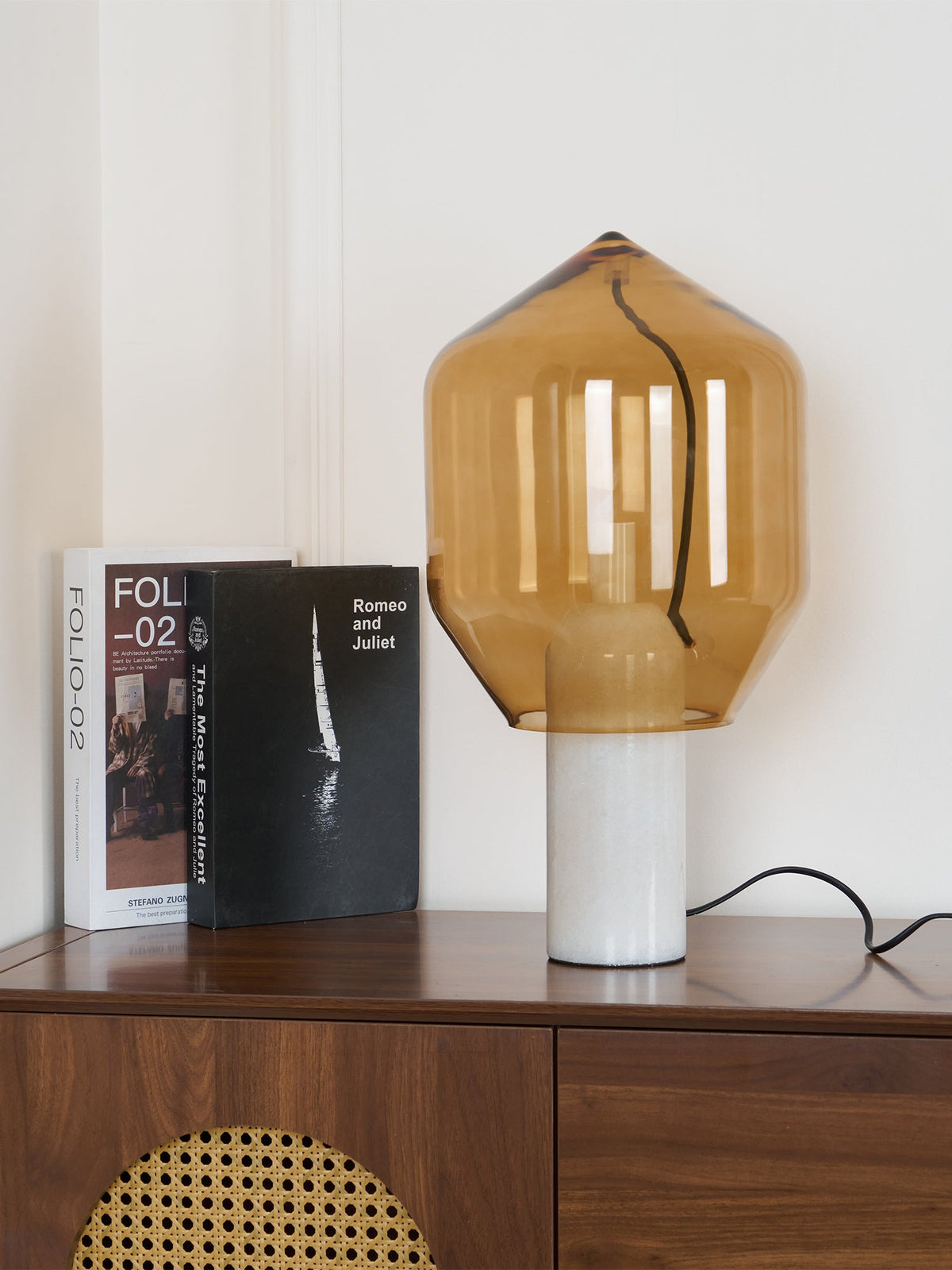An amber-glass Clérachic table lamp on white marble, shown unlit beside two books on a walnut credenza.