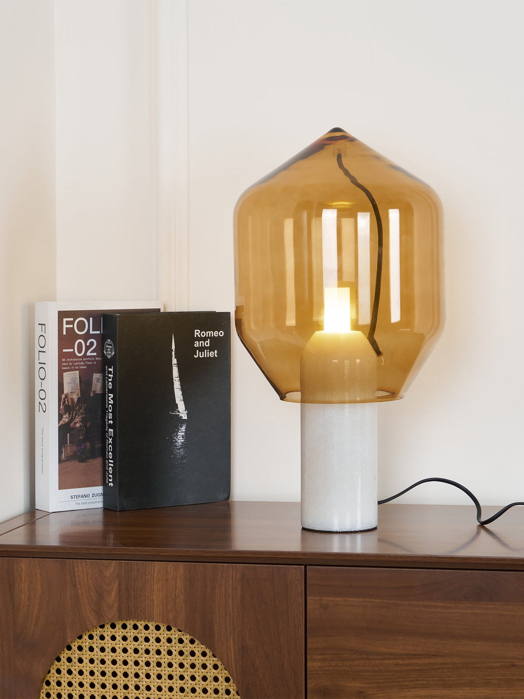 The same amber-glass Clérachic lamp now lit, its bulb glowing warm beside the books on the credenza.