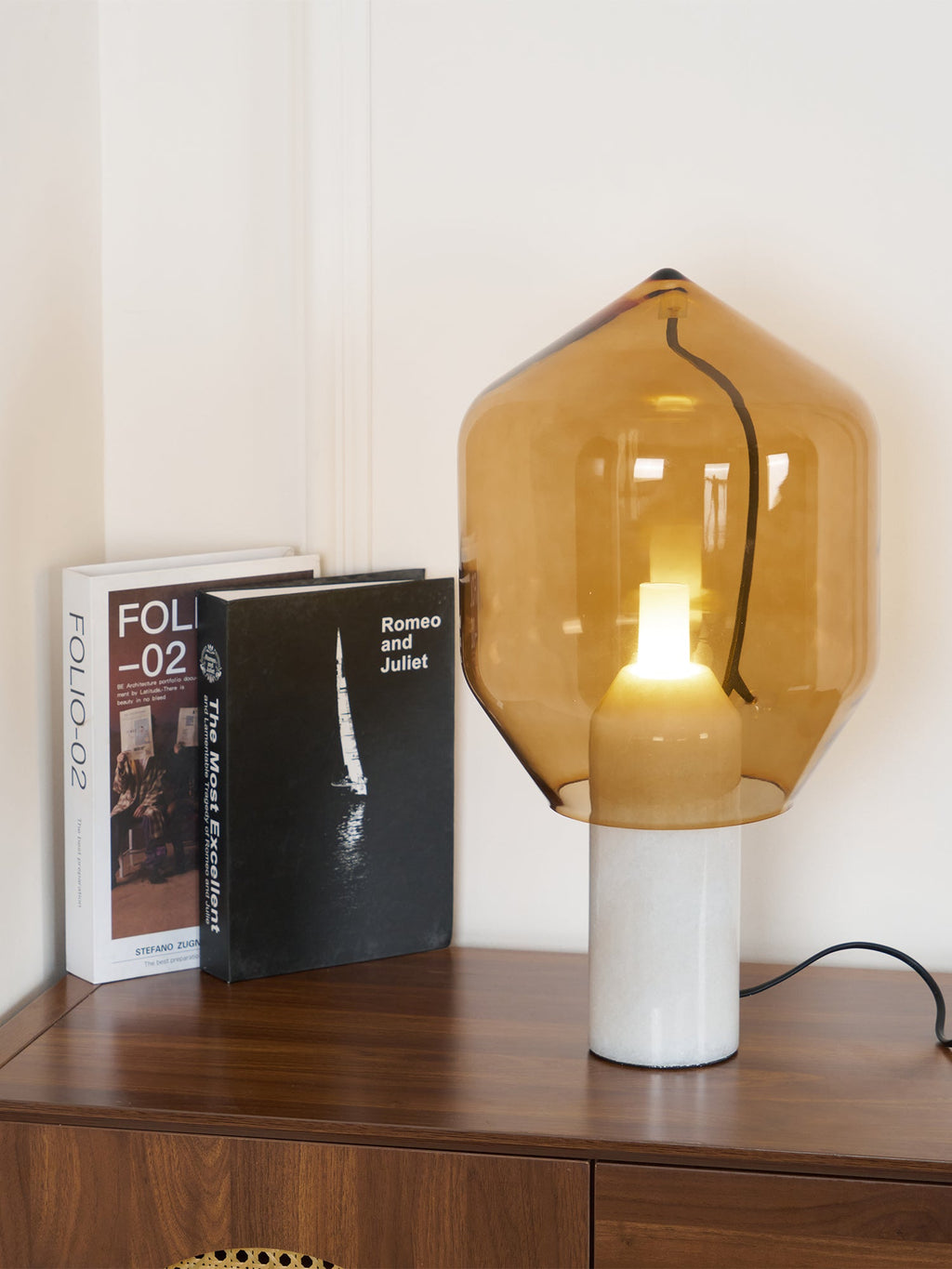The same amber-glass Clérachic lamp cropped closer against the books on the credenza, its cord now hidden from view.