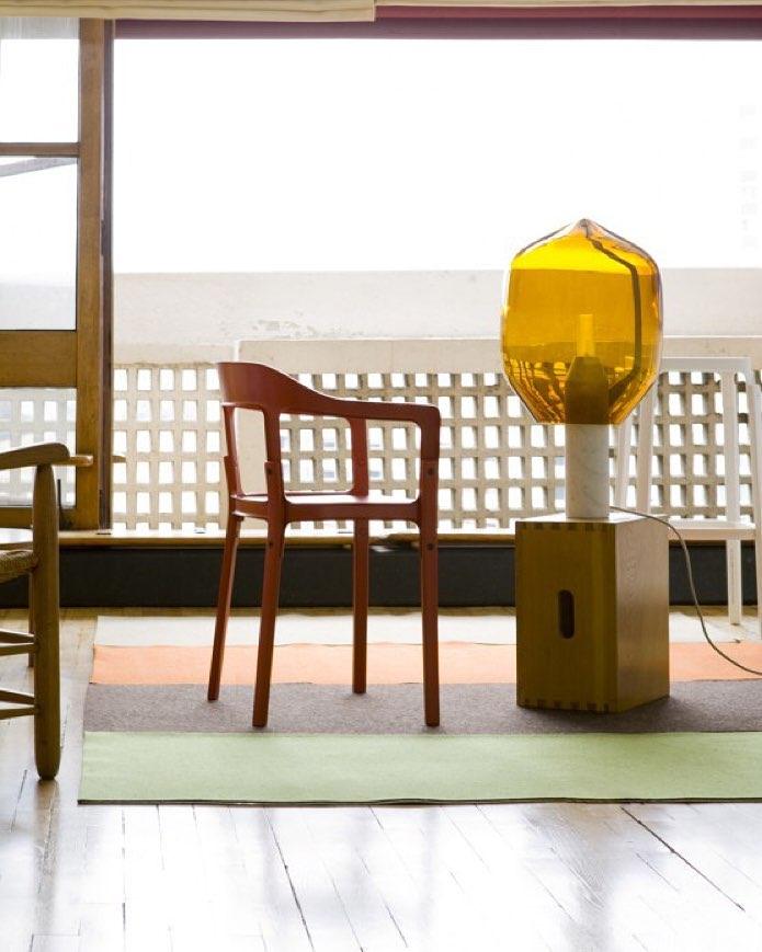 An amber-glass Clérachic lamp on a dark wood plinth, set on a sunlit balcony beside a vintage armchair.
