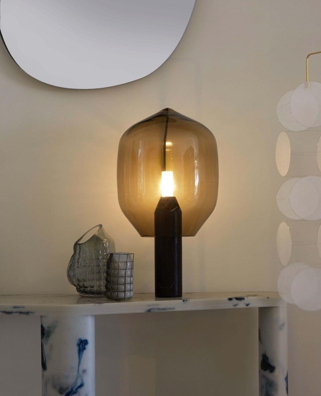An amber-glass Clérachic table lamp lit on a marble console, beside a small vase and a round wall mirror.