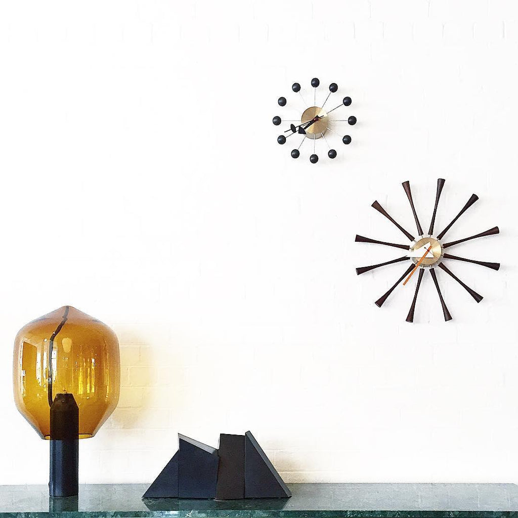 Clérachic's amber-glass lamp on its black base, lit on a glass shelf near two mid-century wall clocks.