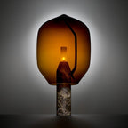 An amber-glass Clérachic lamp on a dark veined marble base, backlit against a charcoal studio background.