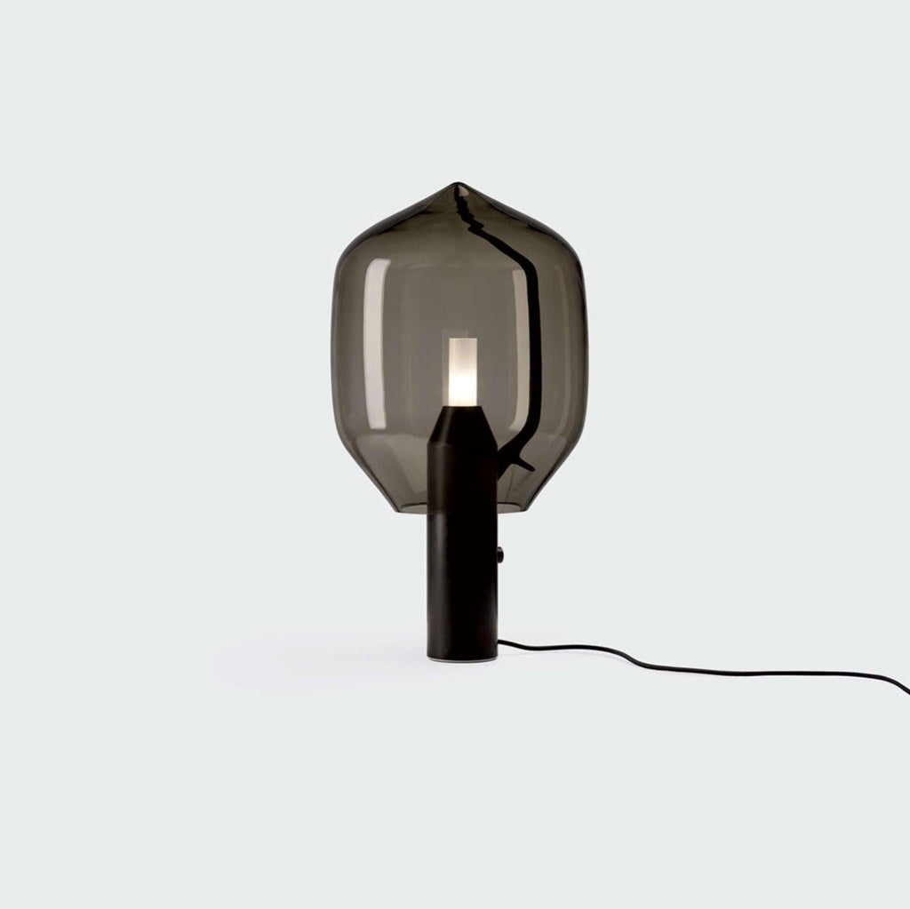Clérachic's smoked-glass lamp on a black base, its cord visible against a pale grey-green backdrop.