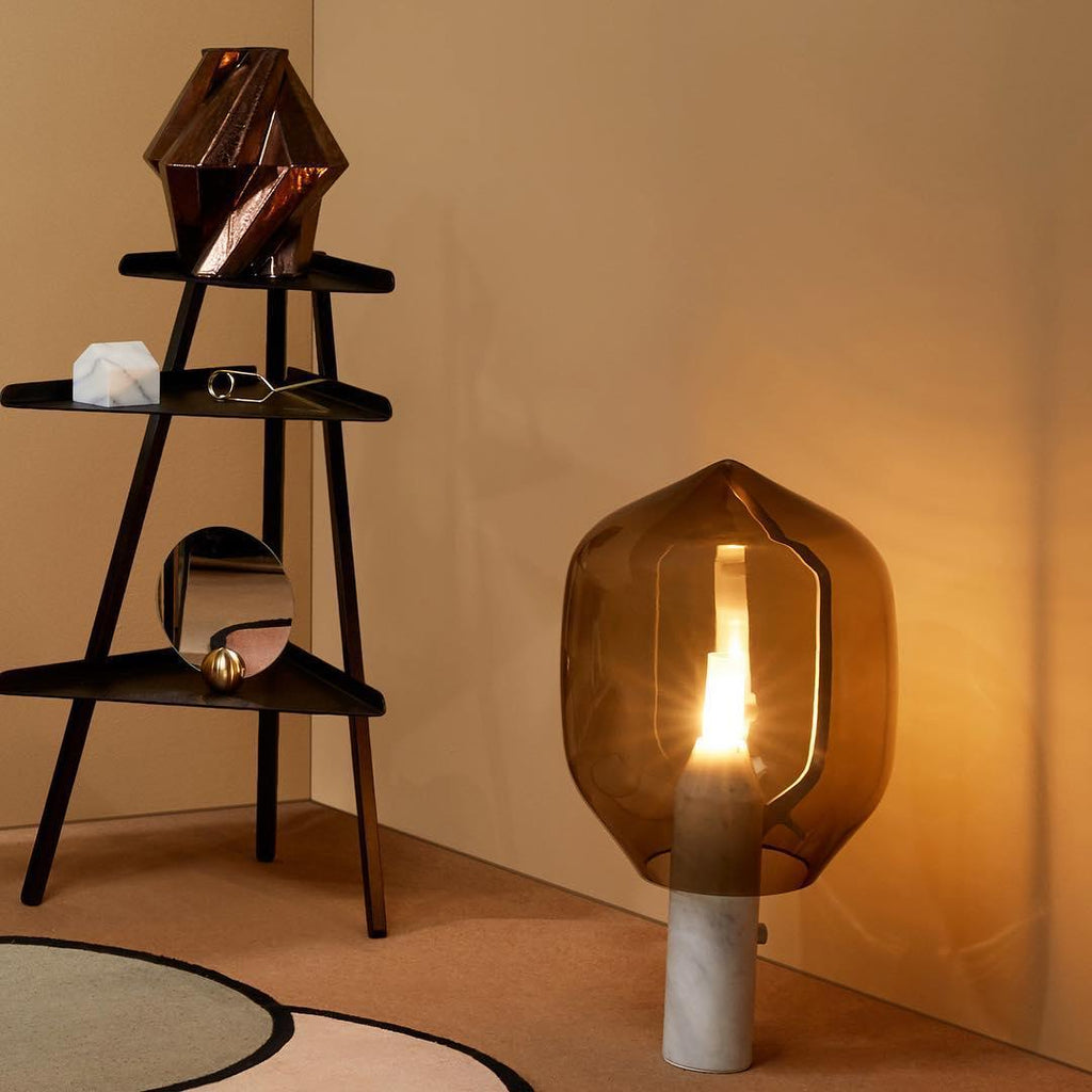A Clérachic table lamp with an olive-toned glass shade and white marble base, glowing in a room corner beside a black ladder shelf.