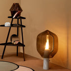 A Clérachic table lamp with an olive-toned glass shade and white marble base, glowing in a room corner beside a black ladder shelf.