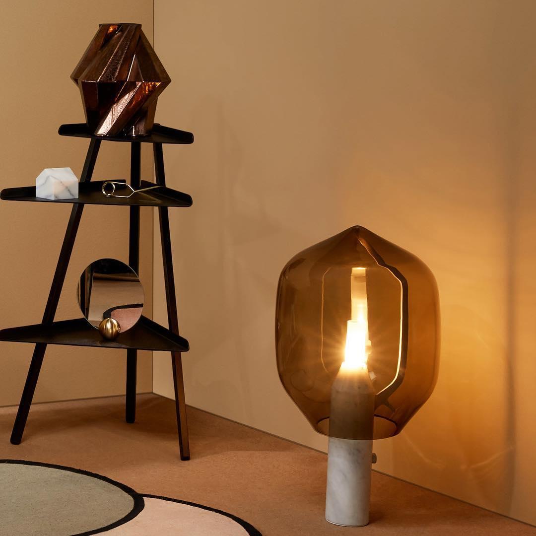 A Clérachic table lamp with an olive-toned glass shade and white marble base, glowing in a room corner beside a black ladder shelf.