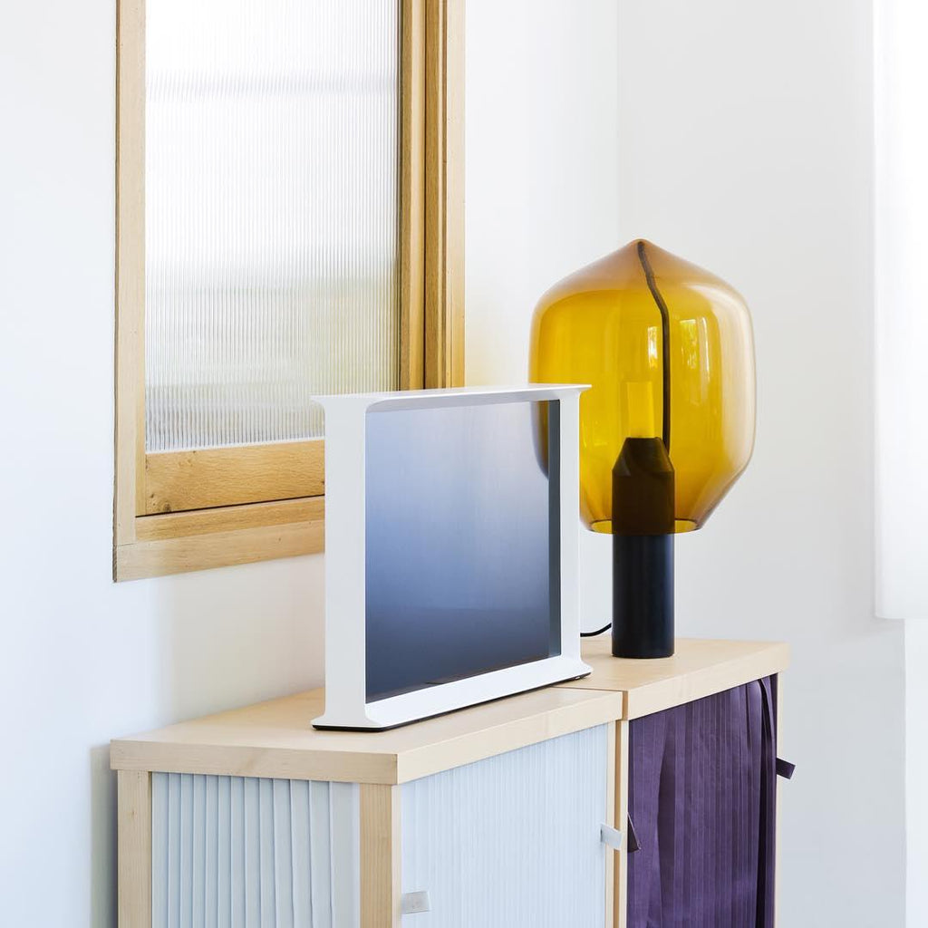 Clérachic's amber-glass lamp lit on a cabinet beside a white flat-screen television and a purple curtain.